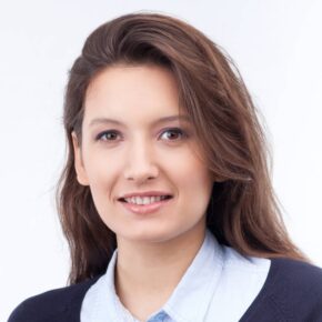 A woman with long brown hair and a bright smile is looking directly at the camera. She is wearing a light blue collared shirt under a dark sweater. The background is plain white, emphasizing her features.