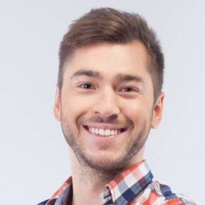 A smiling young man with short brown hair and a beard, wearing a checkered shirt in shades of blue, red, and white, is looking directly at the camera against a light gray background.